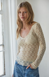 Woman wearing a beige lace top and blue jeans standing in a softly lit room.