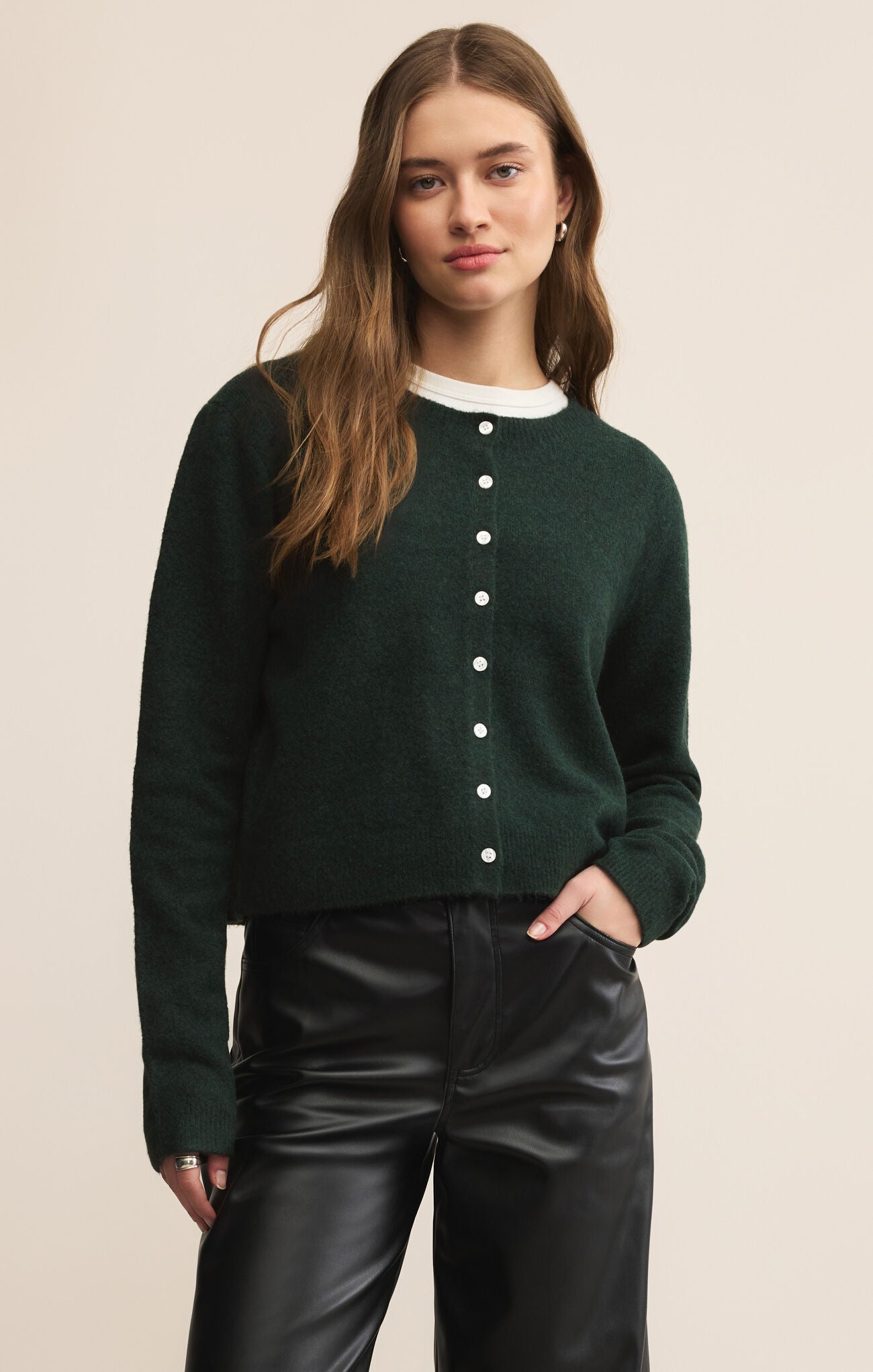 Woman wearing a dark green cardigan with white buttons and black pants on a beige background