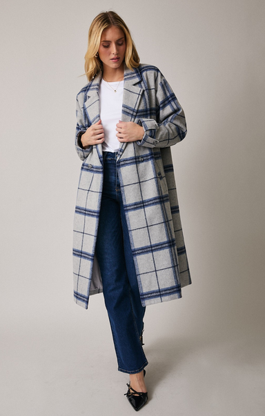 Back view of a woman wearing a long, grey plaid coat with blue windowpane stripes. She has blonde hair, gold hoop earrings, and her hands tucked into the coat pocket