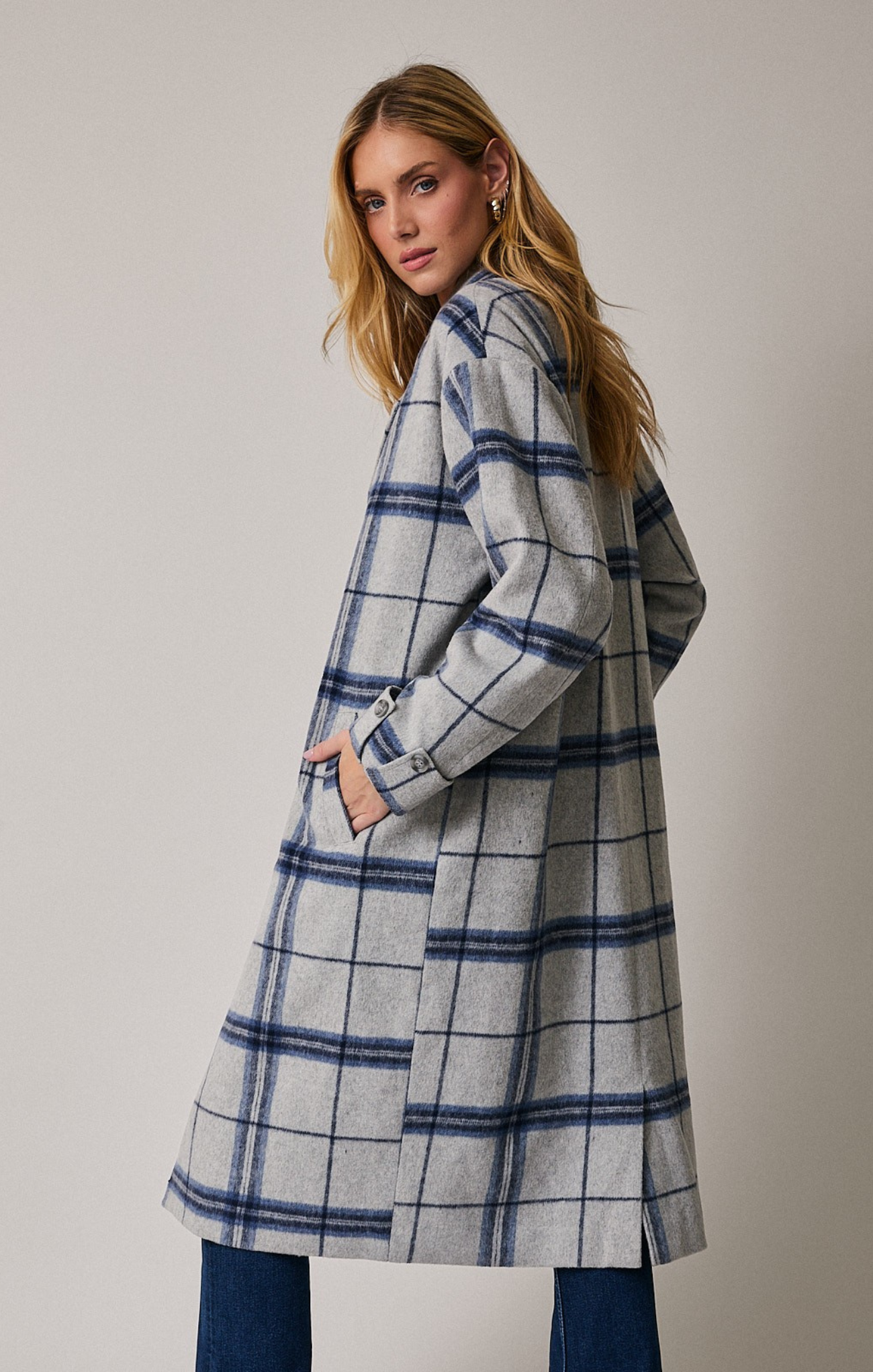 Back view of a woman wearing a long, grey plaid coat with blue windowpane stripes. She has blonde hair, gold hoop earrings, and her hands tucked into the coat pockets