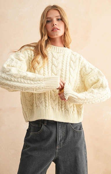 Woman wearing a cream knitted sweater and dark jeans against a beige background