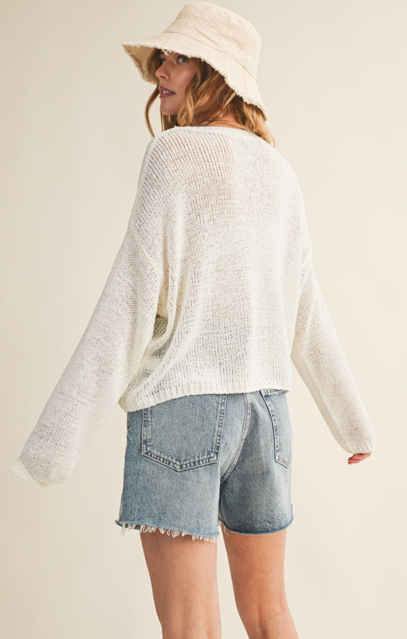 Person wearing a white sweater and denim shorts with a beige hat on a plain background