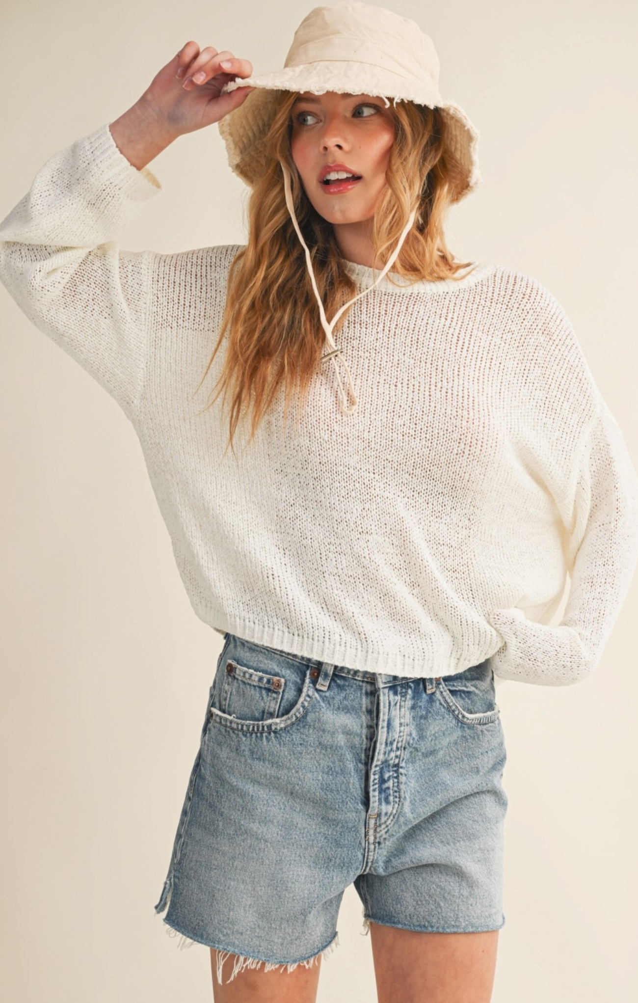 Woman wearing a white sweater and denim shorts with a beige hat on a plain background