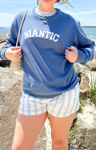 Person wearing a blue sweatshirt with 'Niantic' text and checkered shorts by the ocean.