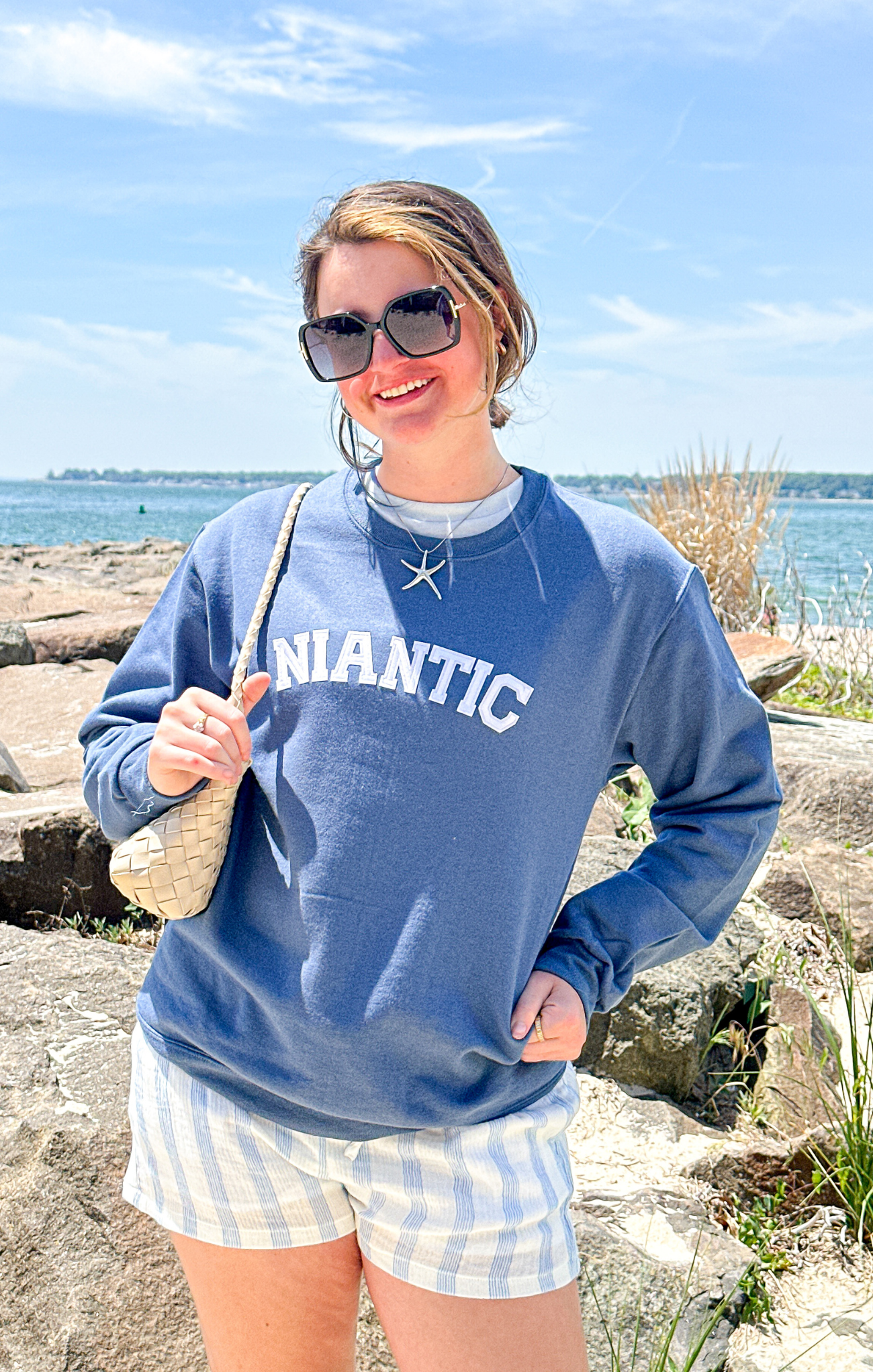 Woman wearing a blue sweatshirt with 'Atlantic' text, sunglasses, and a starfish necklace by the water.