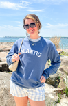 Woman wearing a blue sweatshirt with 'Atlantic' text, sunglasses, and a starfish necklace by the water.