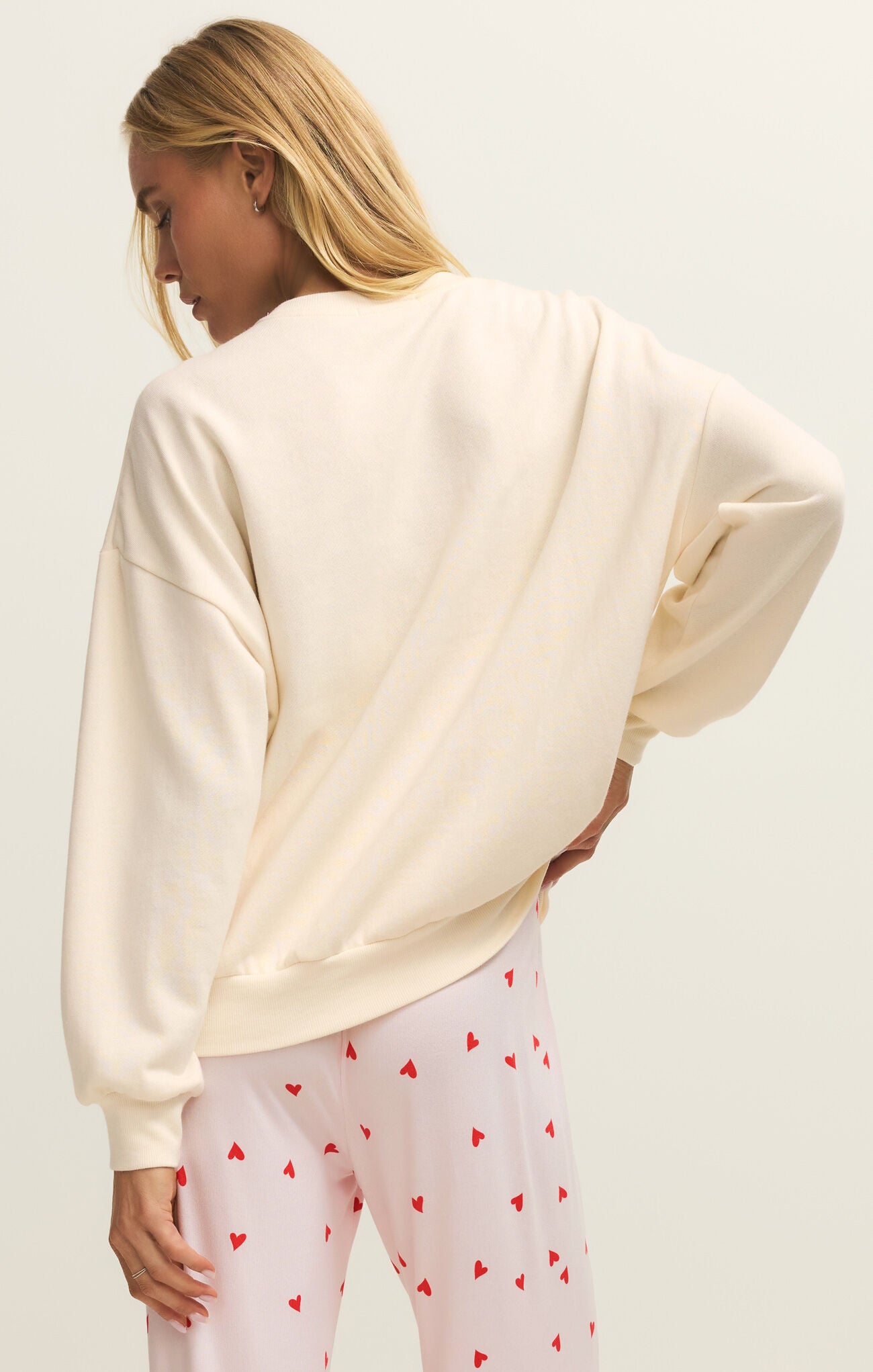 Woman wearing a cream oversized sweater and white pants with red heart patterns on a plain background
