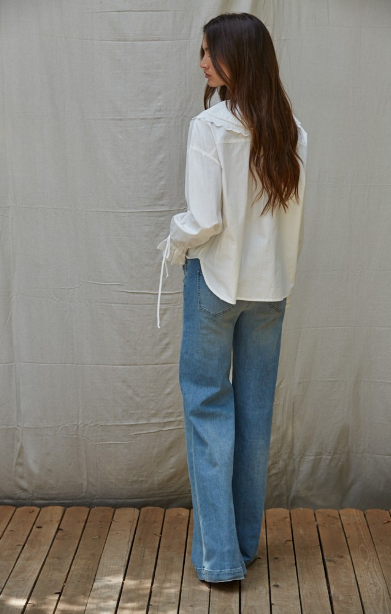 Woman wearing a white blouse and blue jeans standing against a plain wall.