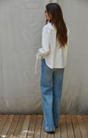 Woman wearing a white blouse and blue jeans standing against a plain wall.