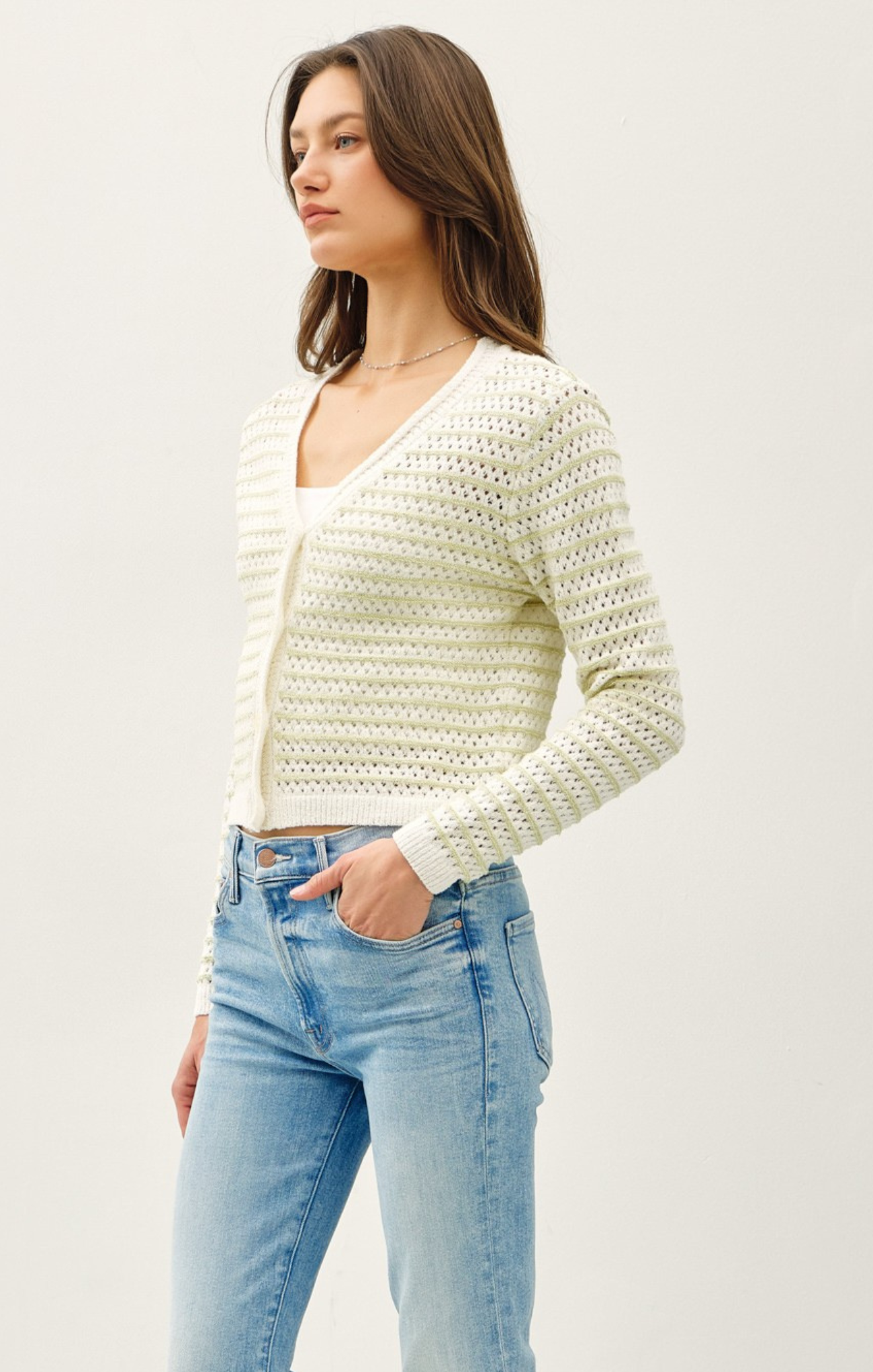 Woman wearing a light-colored knit cardigan over a white top with blue jeans on a plain background