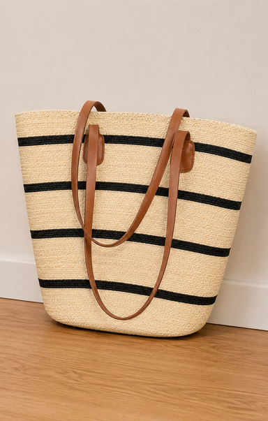 Straw tote bag with black stripes and brown handles on a wooden floor.