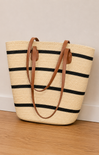 Straw tote bag with black stripes and brown handles on a wooden floor.