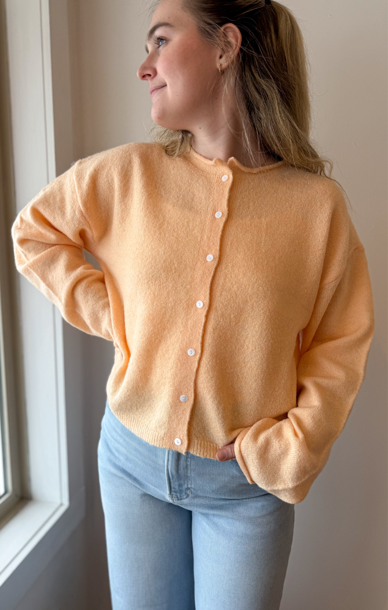 Woman wearing a peach cardigan and light blue jeans indoors.