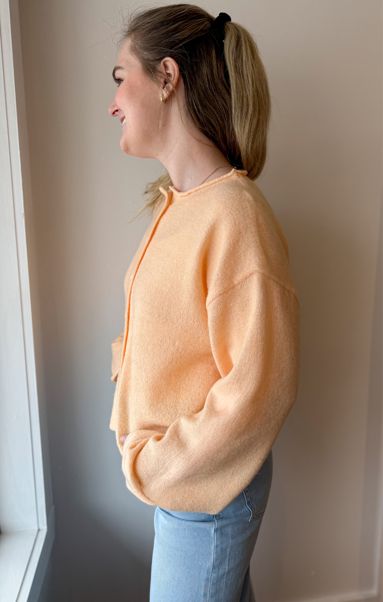 Woman wearing a peach cardigan and blue jeans standing against a plain wall.