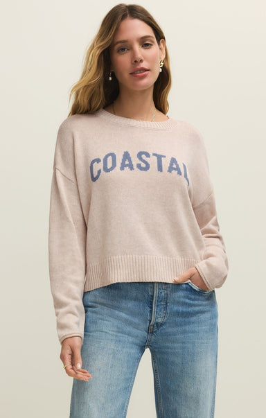 Woman wearing a beige sweater with 'COASTAL' text and blue jeans on a plain background
