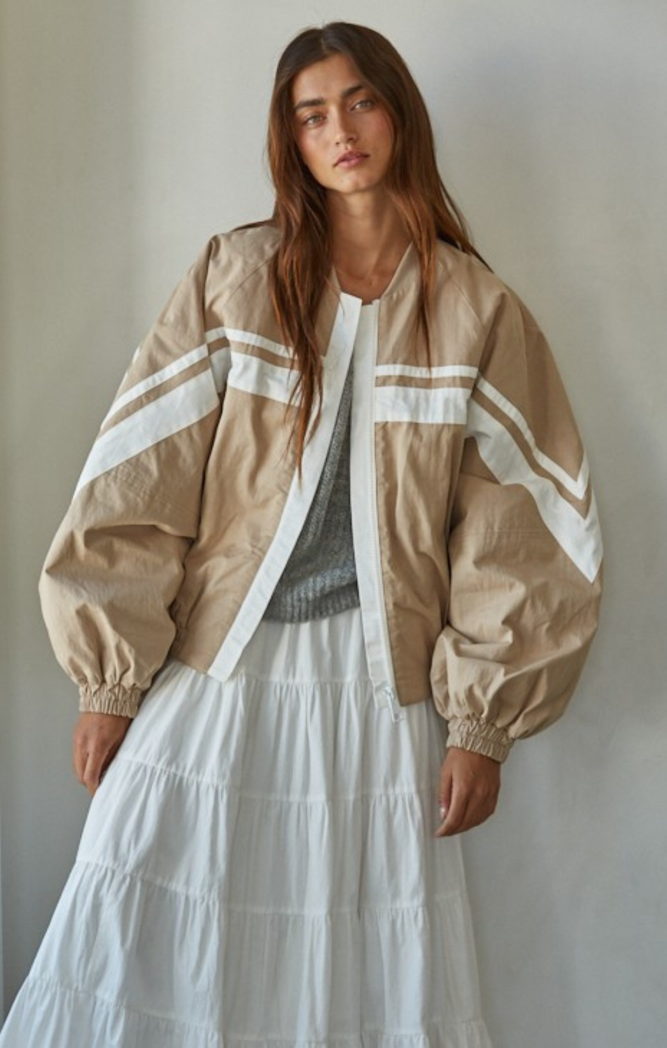 Woman wearing a beige jacket with white stripes and a white skirt against a plain background