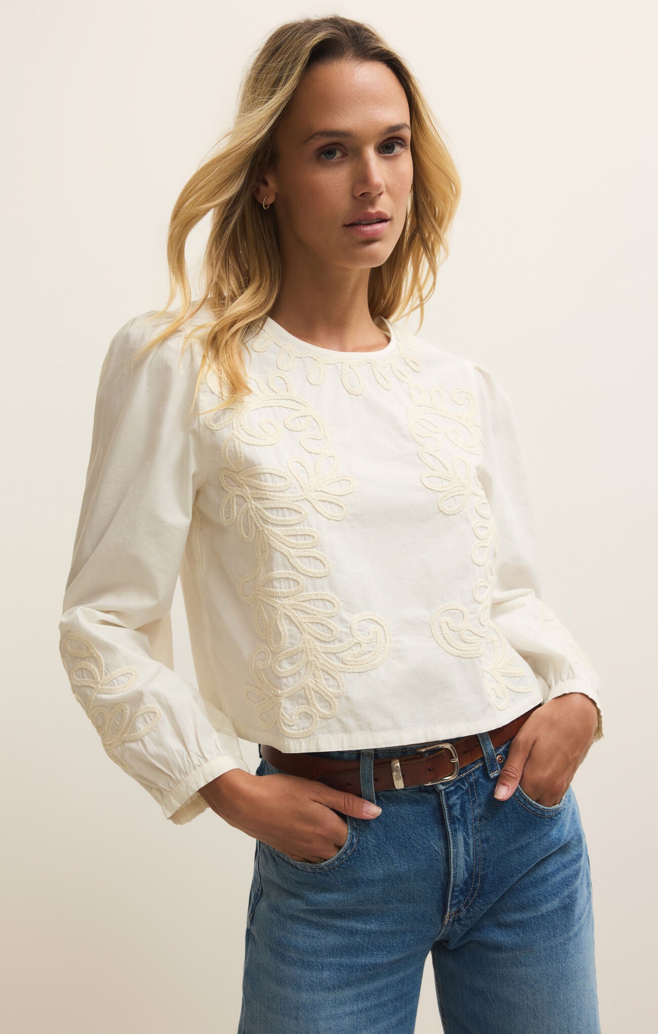 Woman wearing a white blouse with lace details and blue jeans against a plain background