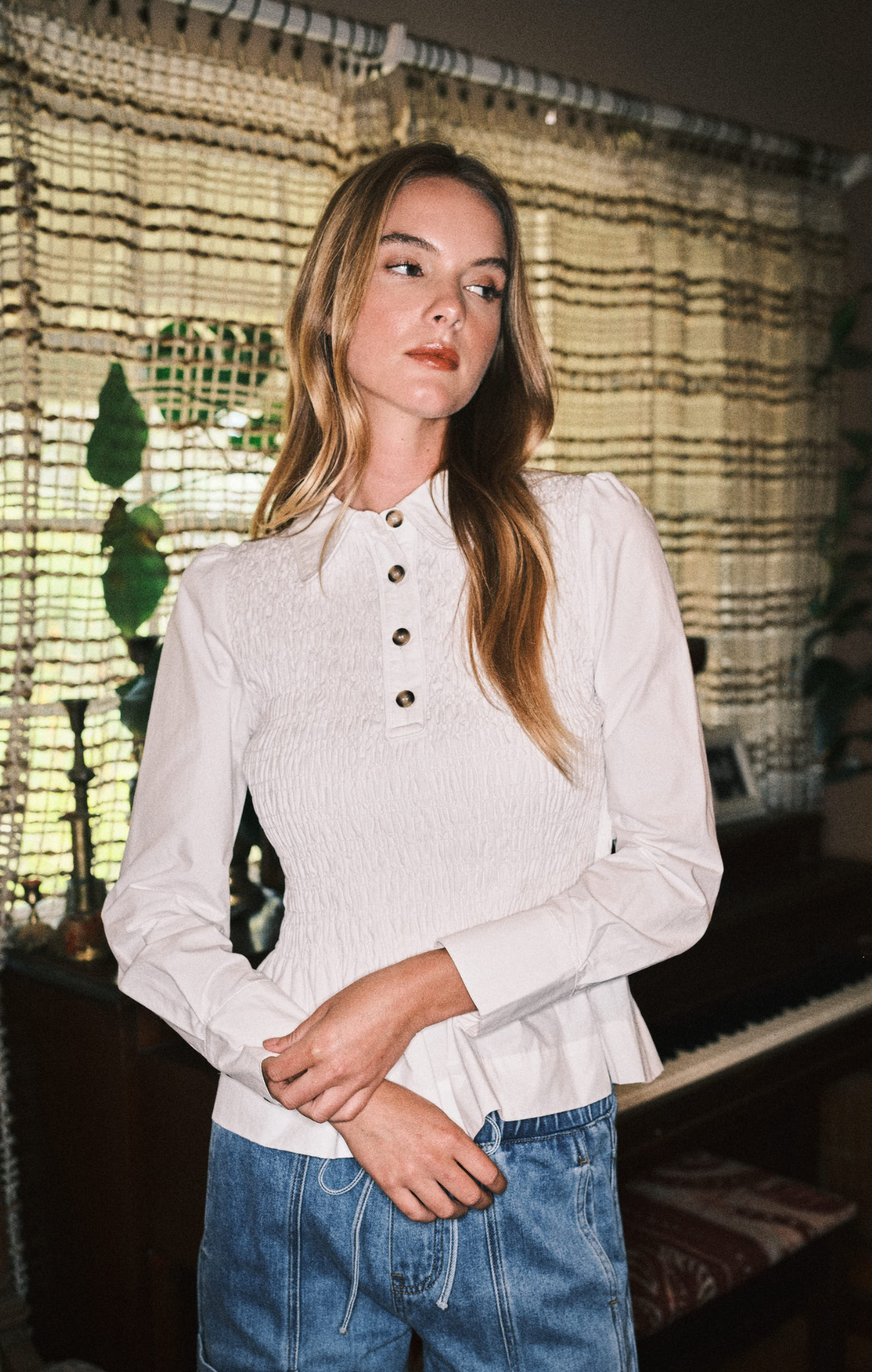 Woman wearing a white blouse with long sleeves and blue jeans in a room with decorative curtains.