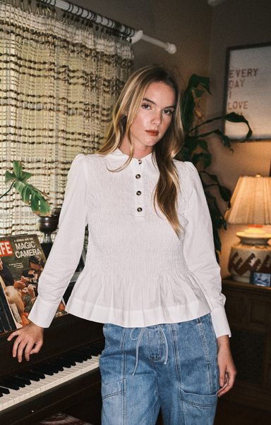 Woman wearing a white blouse with long sleeves and blue jeans in a room with decorative curtains.