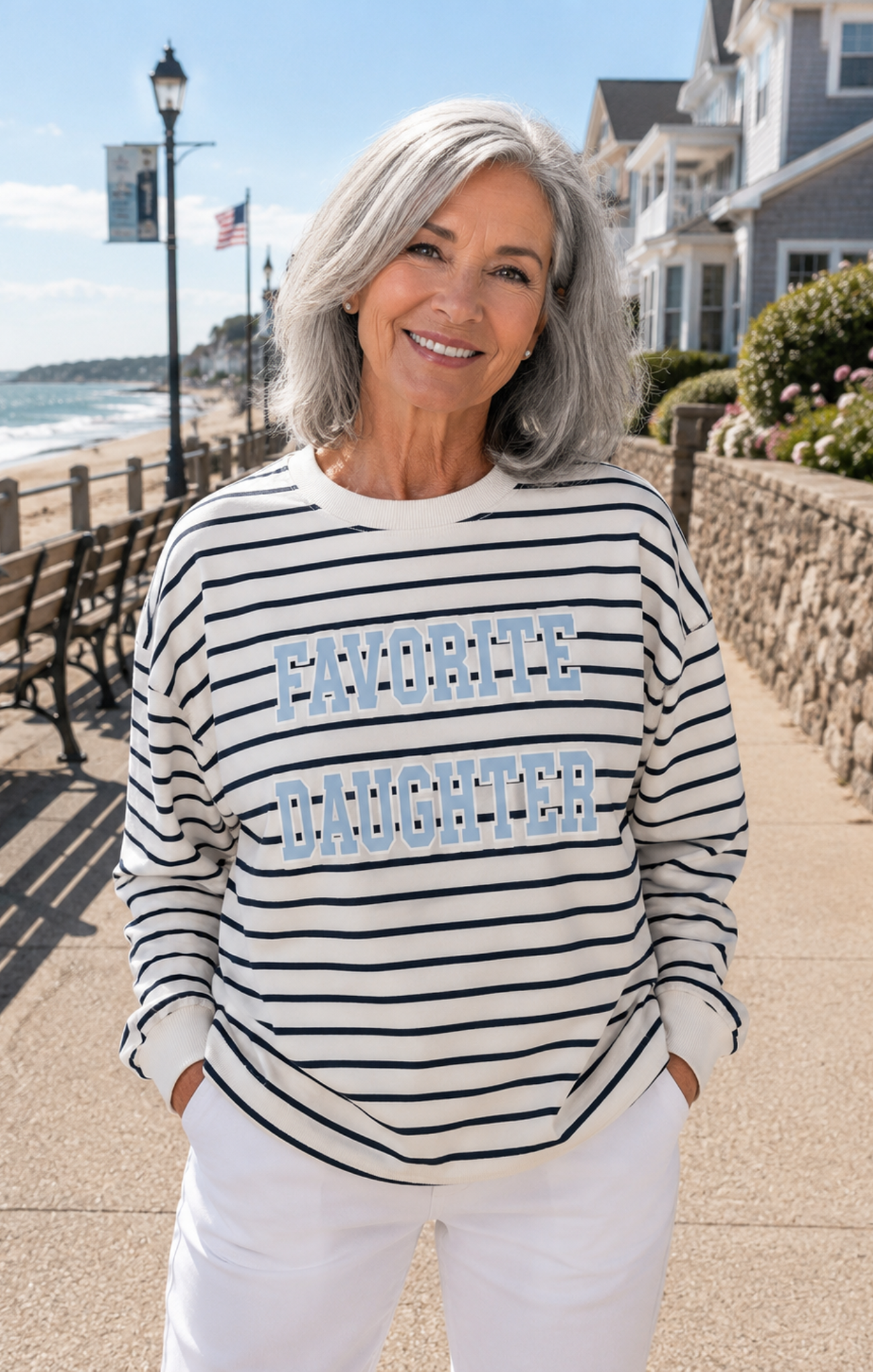 Woman wearing a striped sweater with 'Favorite Daughter' text on a beachside boardwalk.