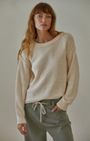 Woman wearing a beige sweater and green pants against a plain background