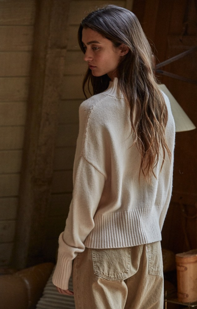 Woman wearing a cream sweater and beige pants in a warm-toned room.
