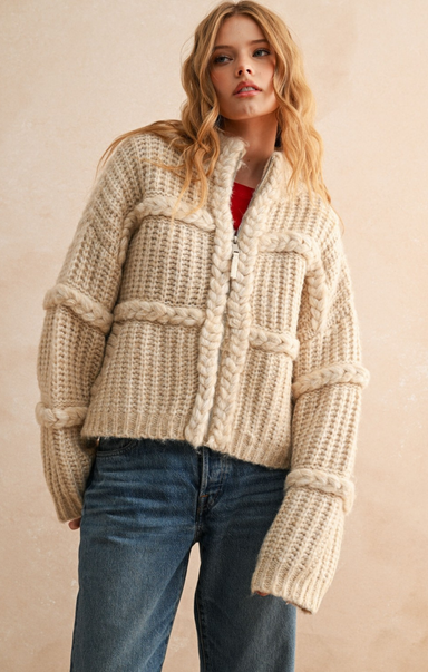 Woman wearing a chunky knit beige sweater and jeans against a plain background