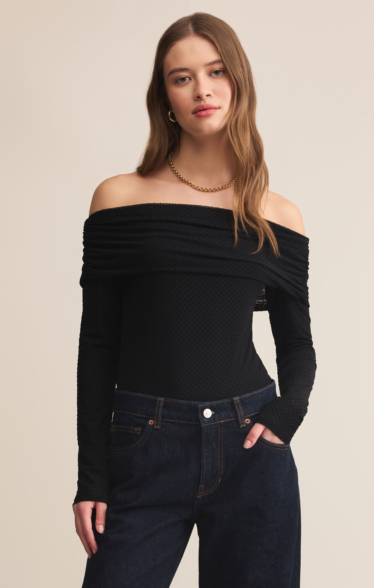 Woman wearing a black off-shoulder top and dark jeans on a beige background