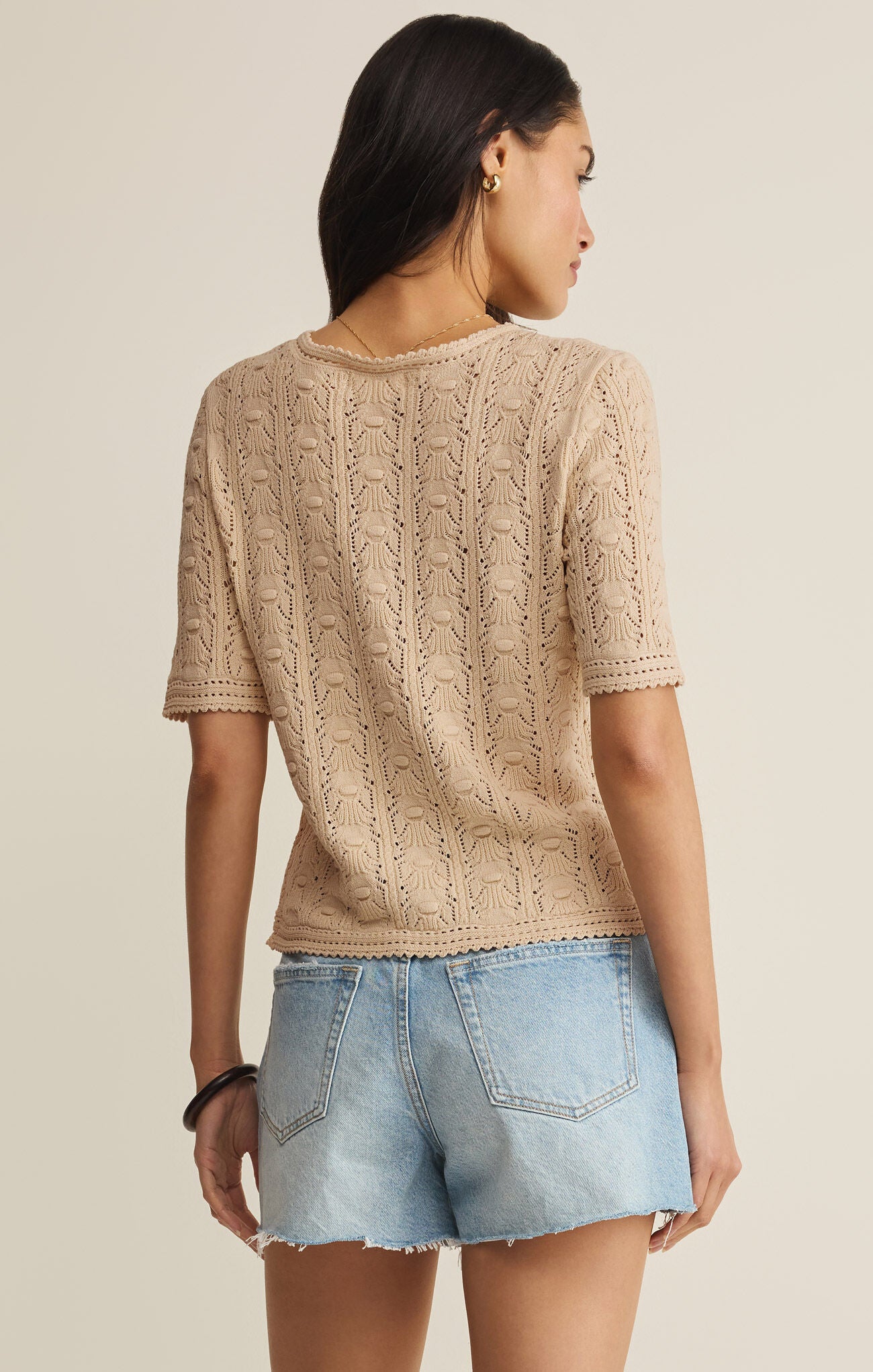 Woman wearing a beige knitted top and denim shorts on a neutral background
