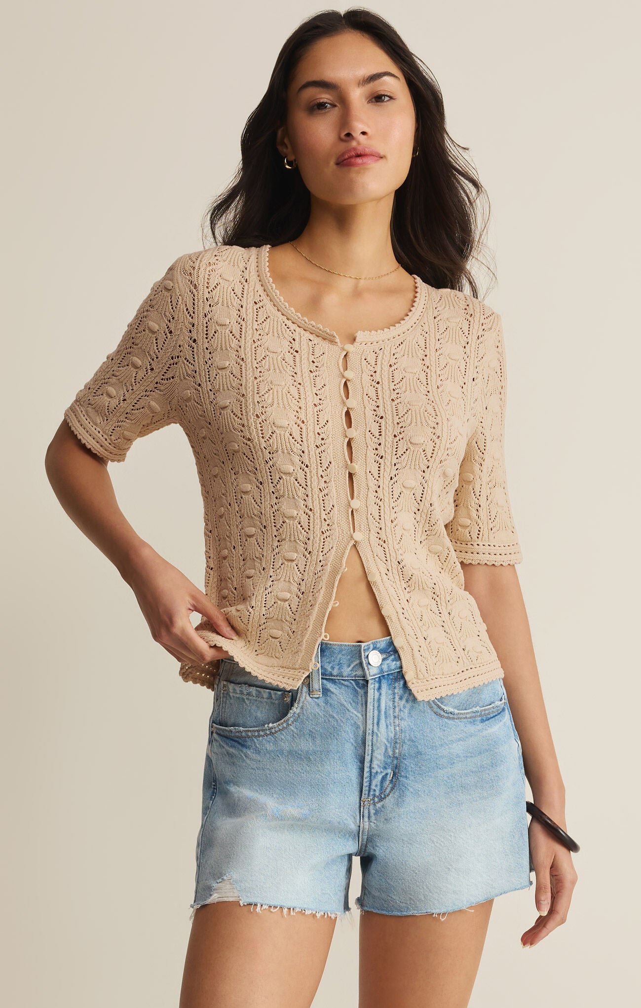 Woman wearing a beige lace cardigan and denim shorts on a plain background