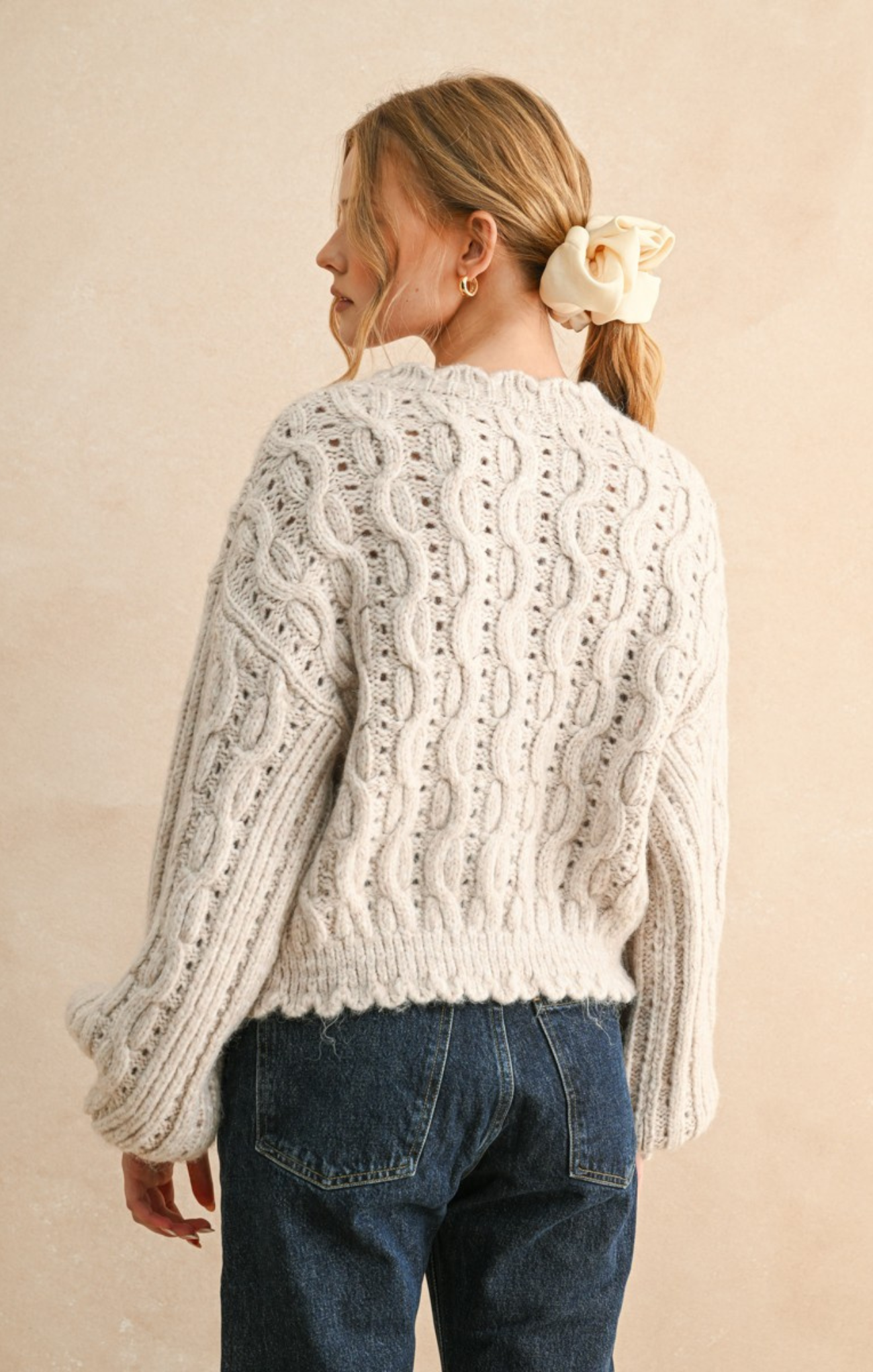 Woman wearing a white cable knit sweater and blue jeans against a beige background