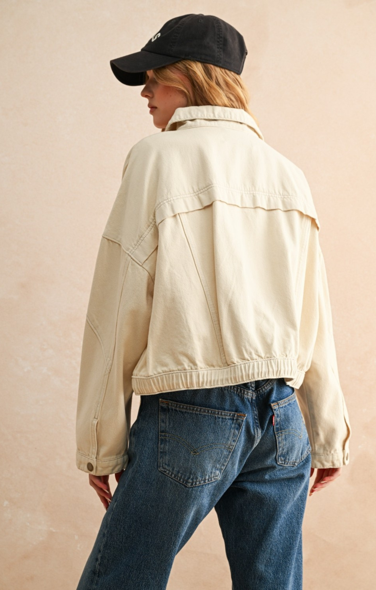 Person wearing a beige jacket and blue jeans with a black cap on a beige background