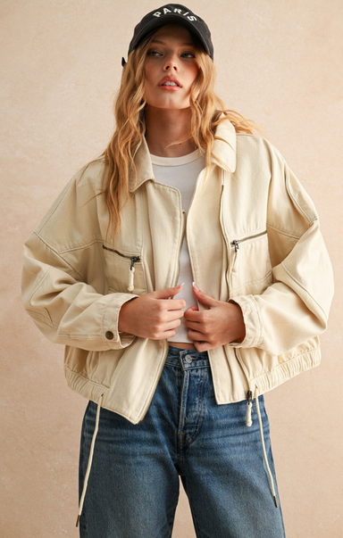 Woman wearing a beige jacket, white shirt, and blue jeans with a black cap on a beige background