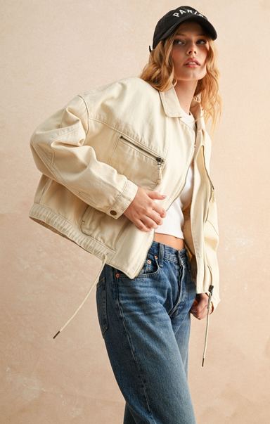 Woman wearing a beige jacket, white shirt, and blue jeans against a beige background