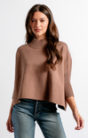A woman modeling a taupe brown knit poncho sweater with a ribbed mock neck and three-quarter sleeves, paired with blue jeans, against a plain white studio background.
