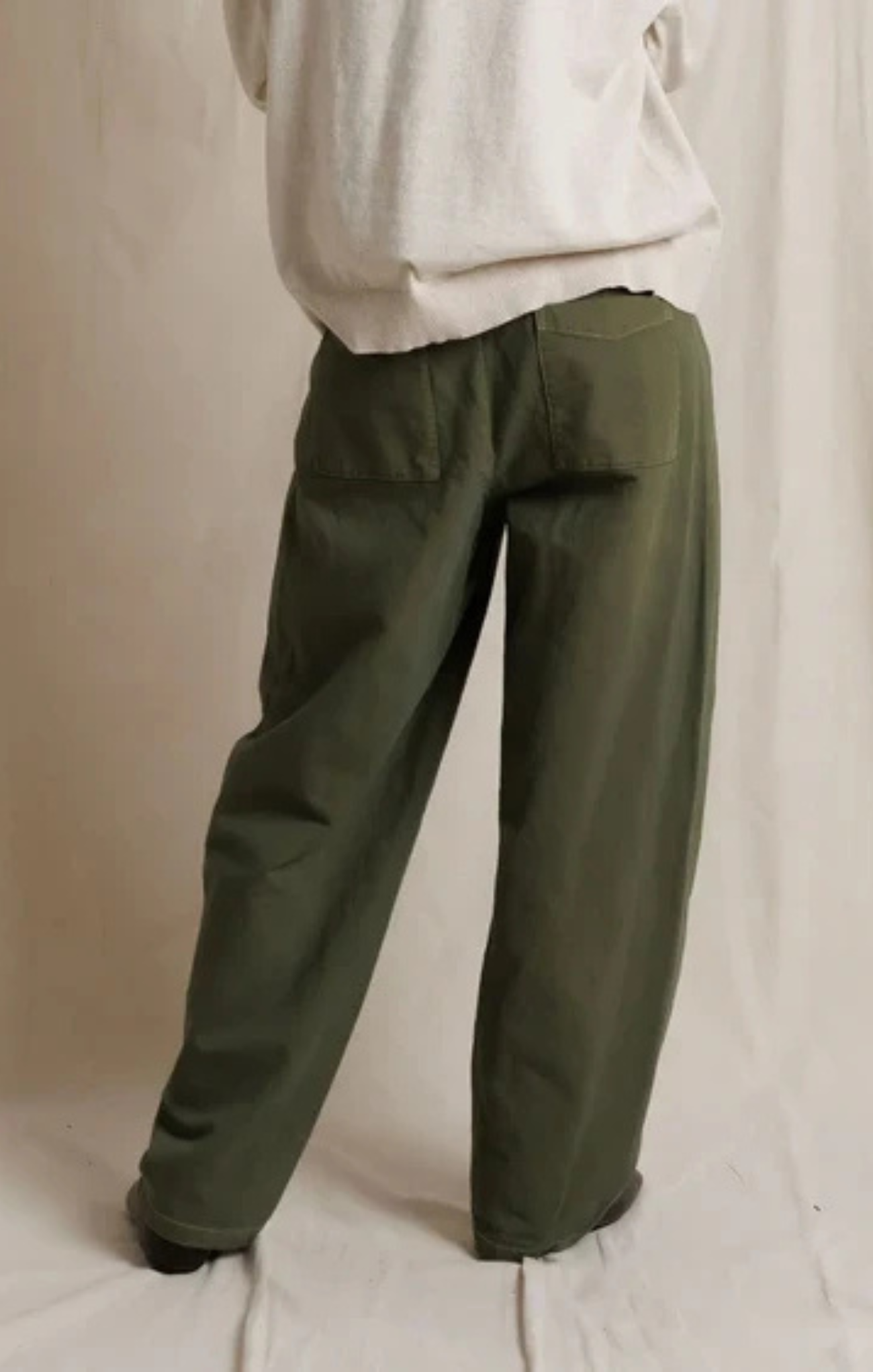 A person shown from the back wearing loose, wide-leg olive green pants and a light cream top, standing against a draped off-white fabric backdrop.
