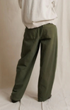 A person shown from the back wearing loose, wide-leg olive green pants and a light cream top, standing against a draped off-white fabric backdrop.