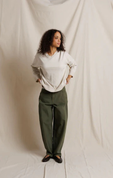 A woman stands against a draped off-white fabric backdrop, wearing an oversized off-white long-sleeve top and dark olive green wide-leg trousers with black shoes.