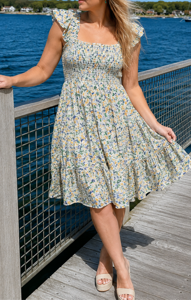 Woman in a floral dress standing on a dock by a body of water.