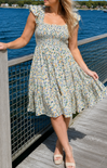 Woman in a floral dress standing on a dock by a body of water.