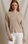 A woman wearing a beige ribbed knit sweater and white pants, standing against a neutral draped fabric backdrop.