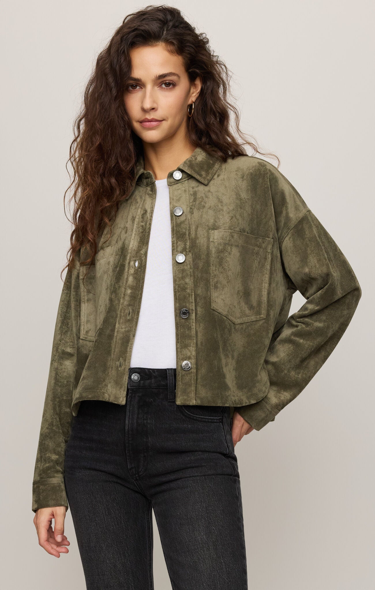 Woman wearing a green suede jacket over a white shirt and black pants on a plain background