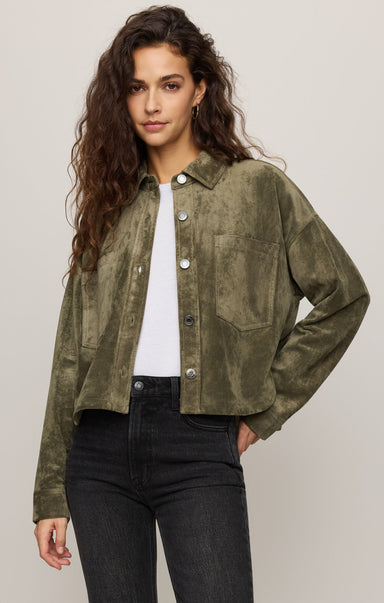 Woman wearing a green suede jacket over a white shirt and black pants on a plain background