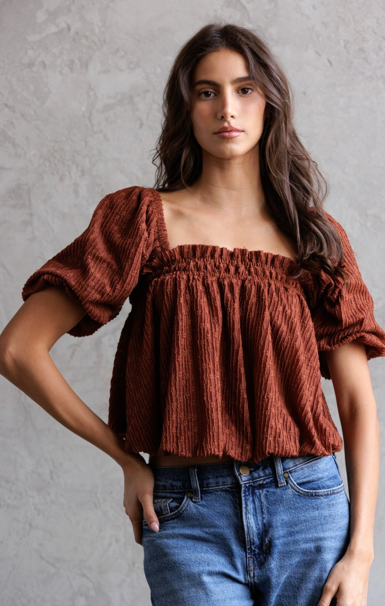 Woman wearing a rust-colored blouse with puffed sleeves and blue jeans against a gray background