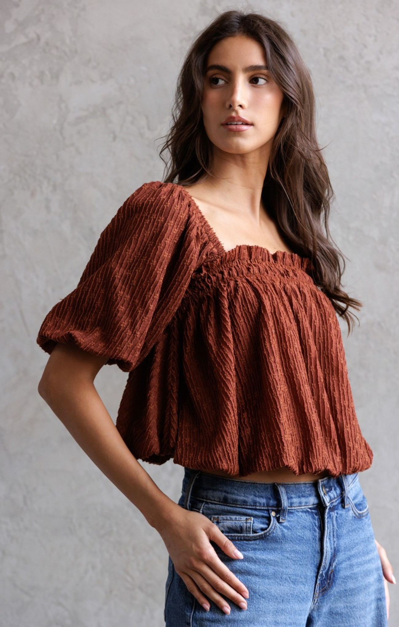 Woman wearing a rust-colored top and blue jeans against a gray background