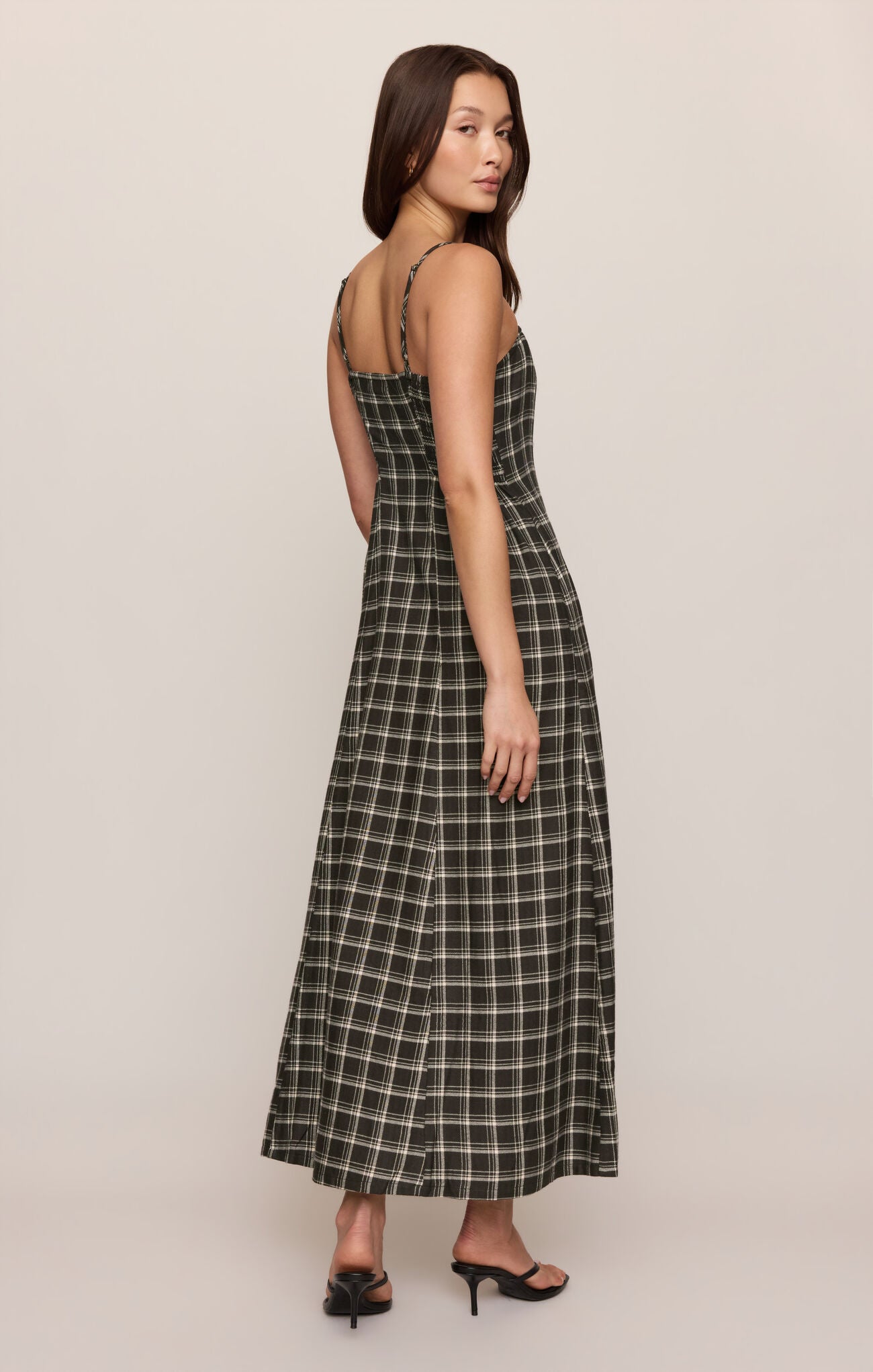 A woman modeling a sleeveless maxi dress with thin straps and a black and white windowpane plaid pattern, shown from a three-quarters back view against a solid light beige studio background.