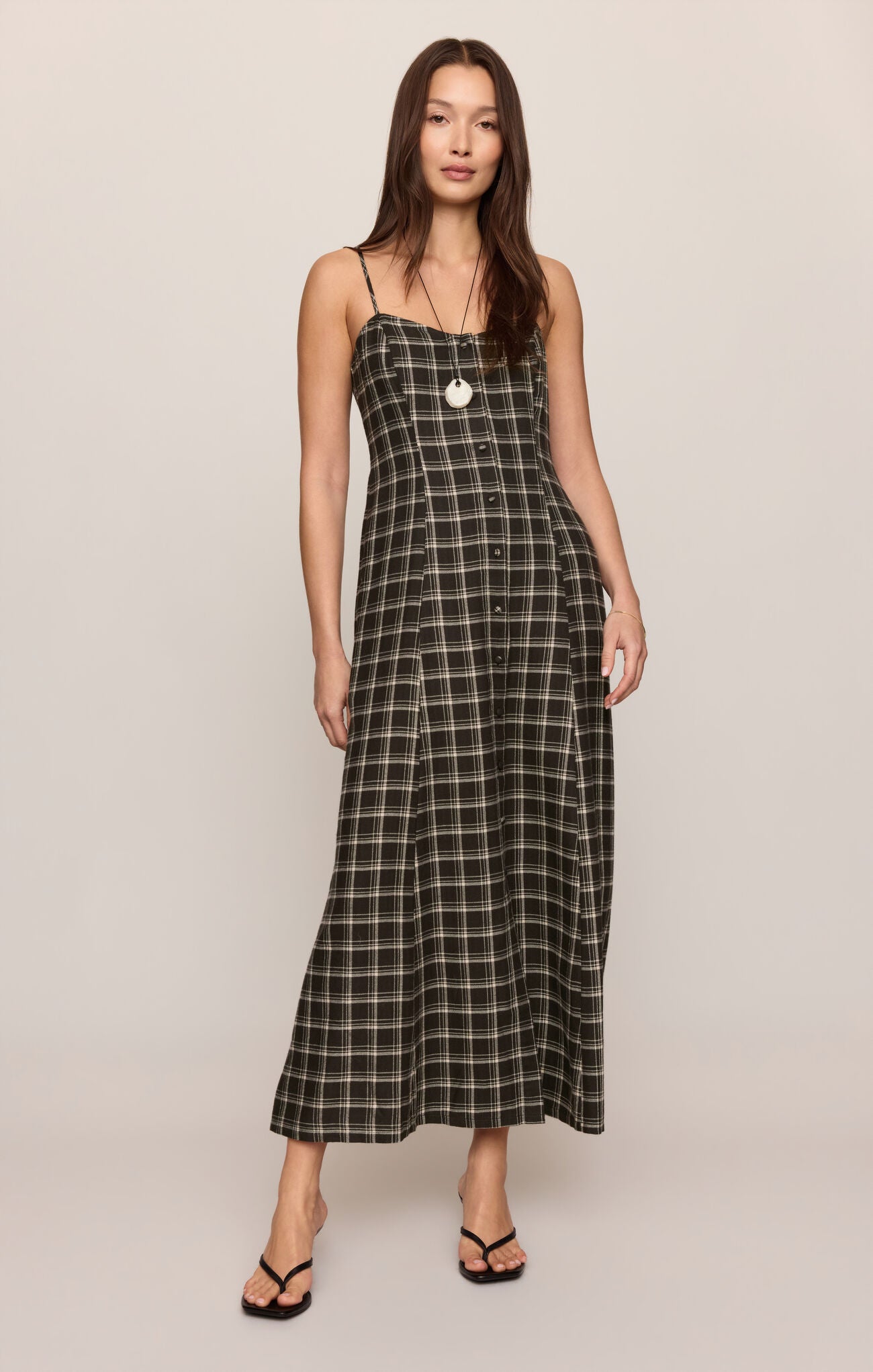 A woman modeling a black and white plaid maxi dress with spaghetti straps and a button-up front, standing against a plain light beige studio background.
