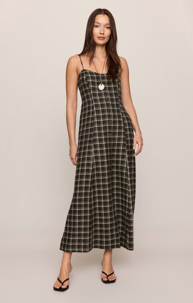 A woman modeling a black and white plaid maxi dress with spaghetti straps and a button-up front, standing against a plain light beige studio background.