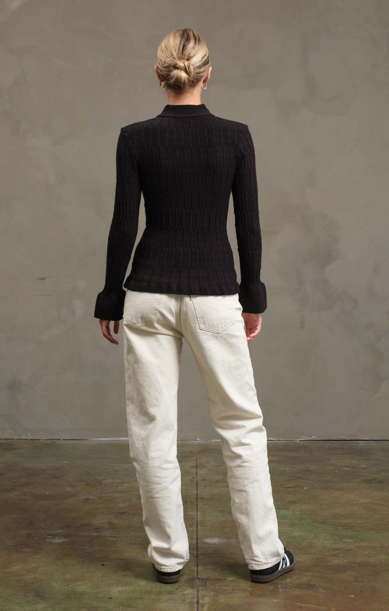 Person wearing a black sweater and white pants against a gray wall.