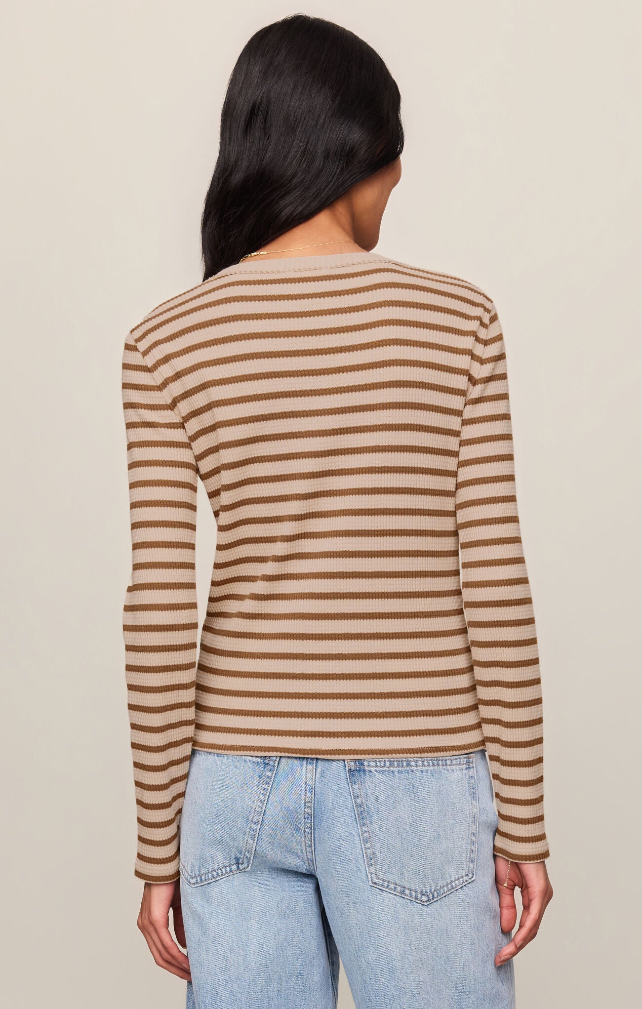 A woman shown from the back wearing a beige and brown horizontally striped long-sleeve knit top with light blue jeans, set against a plain beige studio background.